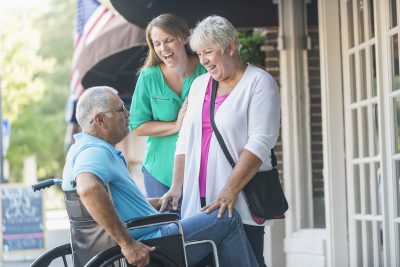A senior couple and their adult daughter on a city sidewalk outside stores. The man is sitting in a wheelchair, looking up at his wife with a serious expression. 